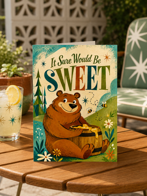 A greeting card featuring a cartoon bear holding a jar of honey with the text 'It Sure Would Be SWEET' in large decorative letters. The card is displayed on a wooden table in an outdoor patio setting with a drink and chair in the background.
