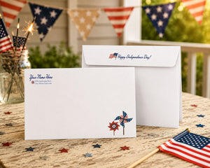 a white envelope with a patriotic design, including a red, white, and blue American flag, along with a pinwheel and stars, placed on a wooden table. There are also American flags and sparklers in the background, suggesting a festive or patriotic setting.