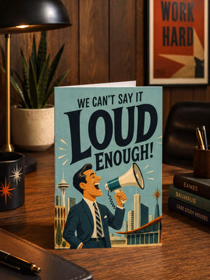 A greeting card with a man in a suit and tie holding a megaphone and shouting "We can't say it loud enough!" The card is placed on a wooden desk with various items such as a lamp, a plant, and books.