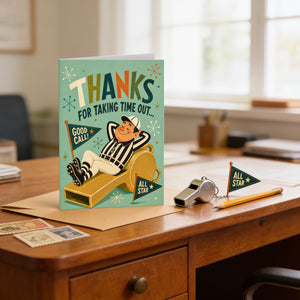 A greeting card with a cartoon illustration of a baseball player sitting on a bench and saying "Thanks for taking time out...", along with a "Good Call" sign and a "All Star" flag. The card is placed on a wooden desk, and there are some postage stamps and a pencil nearby.