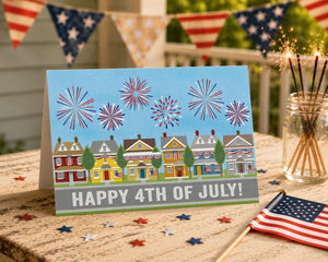 A greeting card with a patriotic design celebrating the 4th of July, featuring fireworks and a row of colorful houses. The card is placed on a wooden table, surrounded by American flags and sparklers.