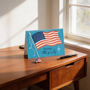 A greeting card with an American flag design and the text "Happy 4th of July" is displayed on a wooden desk, accompanied by a pen and a small American flag pin.