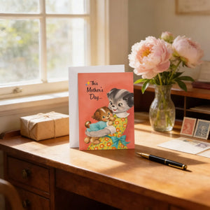 A Mother's Day card with a dog illustration sits on a wooden desk, accompanied by a vase of pink flowers, a gift box, and a pen.