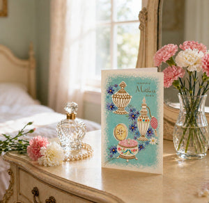 A Mother's Day card with illustrations of perfume bottles and flowers sits on a wooden dresser, accompanied by a vase of pink and white flowers and a crystal perfume bottle.