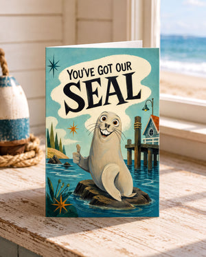 A greeting card featuring a cartoon seal with the text "You've Got Our SEAL" on it, sitting on a rock in a coastal scene.
