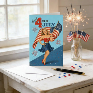 A greeting card celebrating the 4th of July, featuring a vintage illustration of a woman holding an American flag, is displayed on a table with a pen and a sparkler vase.
