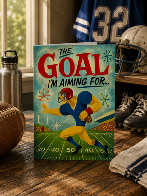 A football-themed greeting card standing on a wooden table, surrounded by sports props like a football, helmet, jersey, and water bottle. The card features retro-style artwork of a football player with the text 'THE GOAL I'M AIMING FOR.