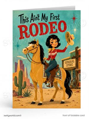 A folded greeting card shown upright on a white background. The front features retro western artwork with the text 'This Ain't My First RODEO' above an illustrated woman riding a tan horse in a desert town scene.