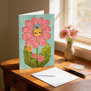 A colorful greeting card featuring a large pink flower with a smiling face is displayed on a wooden desk next to a vase of pink flowers and a pen.