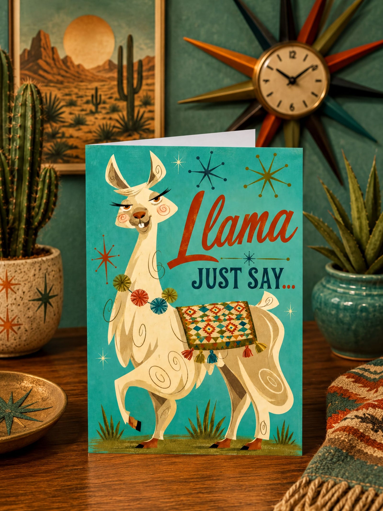 A folded greeting card featuring a stylized cream llama with a colorful geometric saddle blanket against a turquoise background, with the words 'Llama JUST SAY.