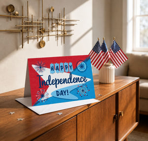 A greeting card with the text "Happy Independence Day!" is displayed on a wooden surface, along with three American flags.