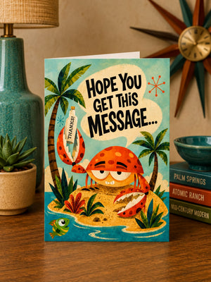 a colorful greeting card with a cartoon crab holding a message, placed on a wooden surface next to a lamp and some decorative items.