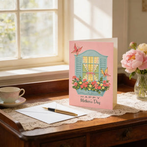 A pink greeting card with a floral design and butterflies sits on a wooden desk, accompanied by a teacup, pen, and a vase of pink flowers.