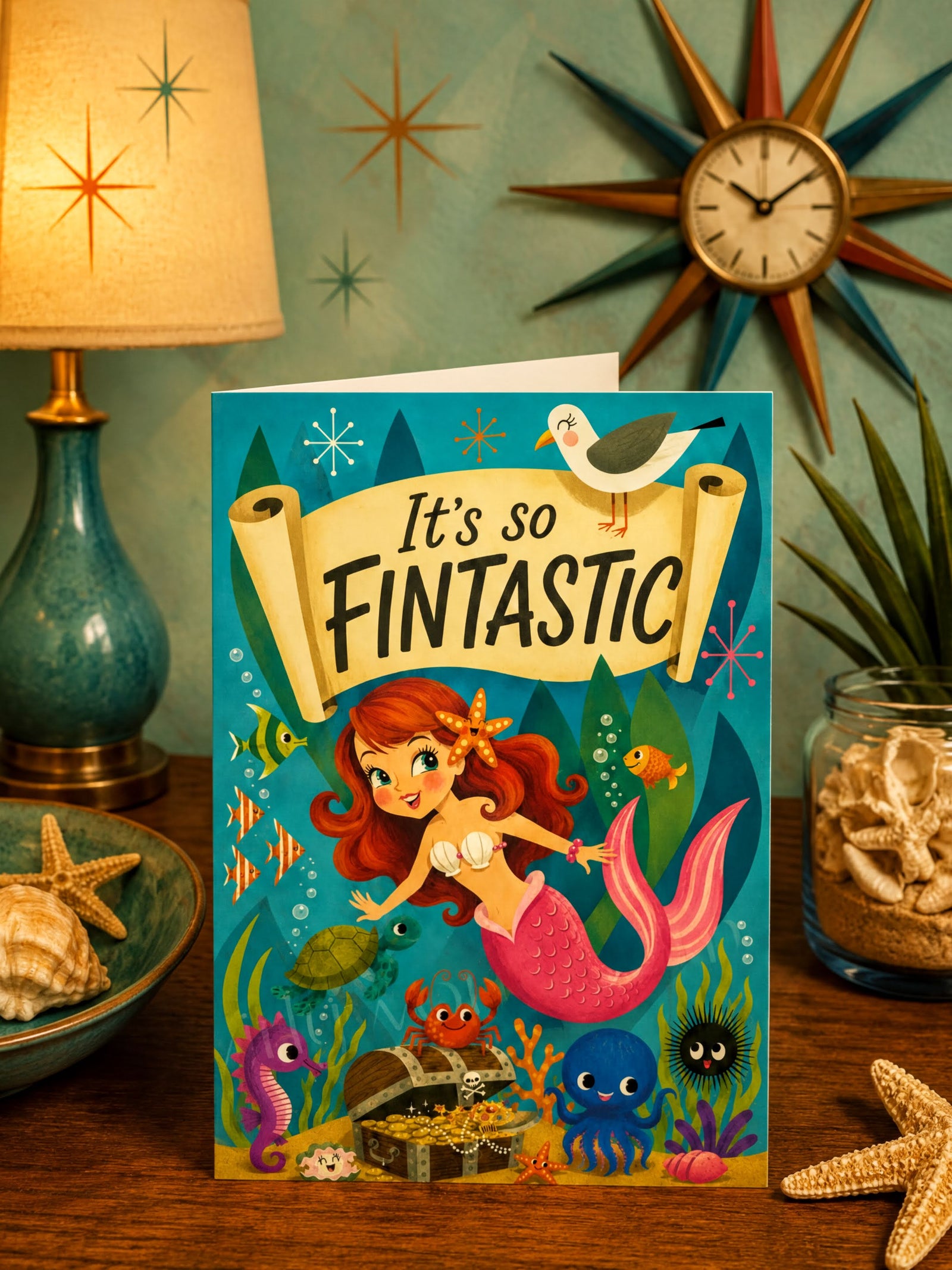 A colorful folded greeting card featuring an underwater mermaid scene with the text 'It's so FINTANTIC' on a scroll banner. The card shows a mermaid with red hair and a pink tail, surrounded by fish, a crab, a treasure chest, a seahorse, a blue octopus, a black sea urchin, and a seagull at the top.