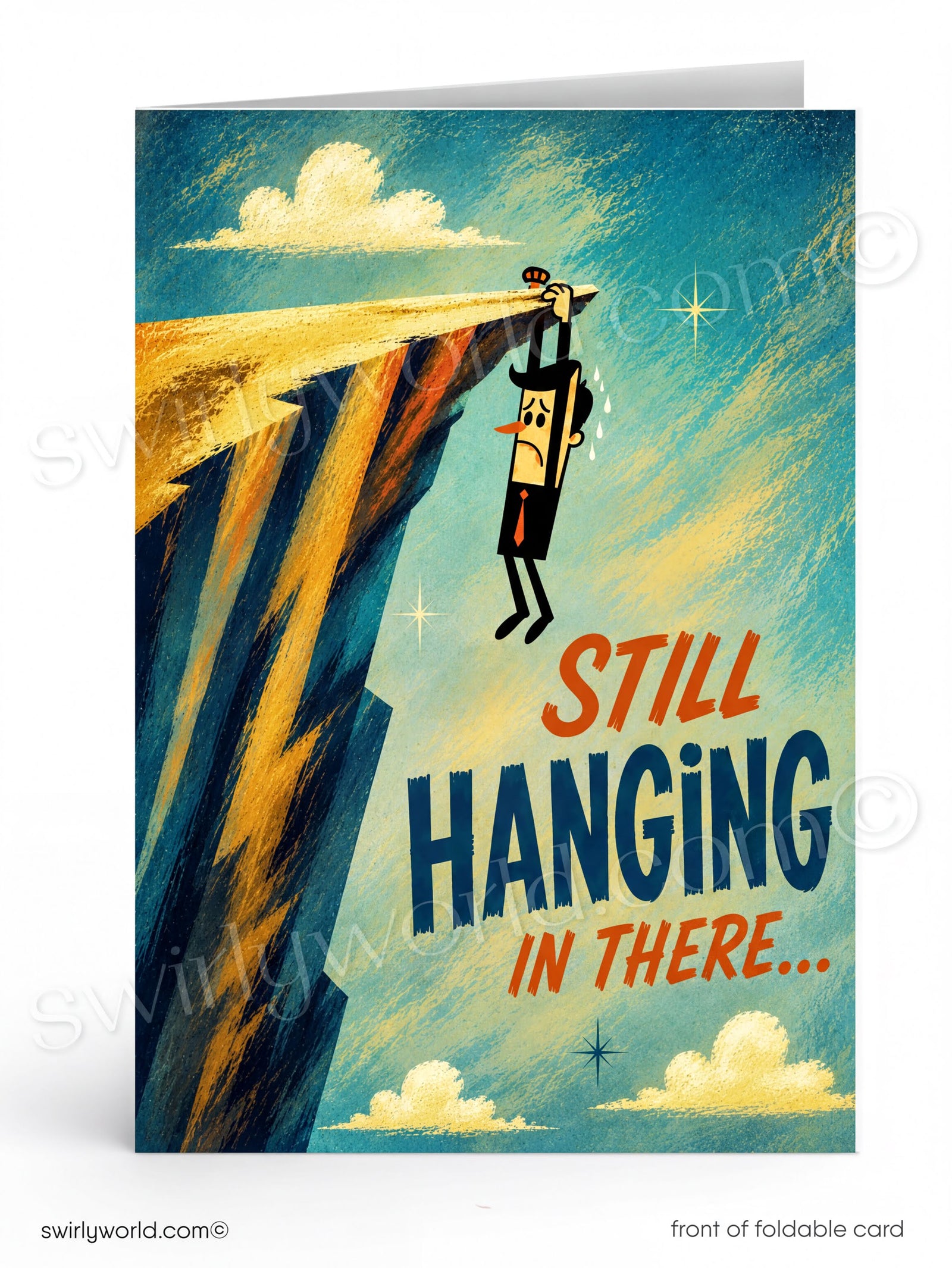 A folded greeting card shown front-on against a white background. The card features an illustrated scene of a small figure hanging from a cliff edge under a blue sky with clouds, and the text 'STILL HANGING IN THERE.