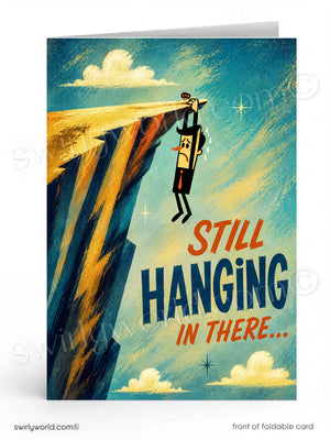 A folded greeting card shown front-on against a white background. The card features an illustrated scene of a small figure hanging from a cliff edge under a blue sky with clouds, and the text 'STILL HANGING IN THERE.