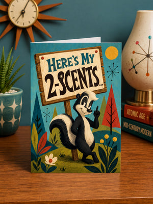 a greeting card with a cartoon skunk holding a sign that says "Here's My 2-Scent..." The card is placed on a wooden surface, with a clock and a lamp visible in the background.