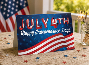 A July 4th greeting card with the text "July 4th Happy Independence Day!" displayed prominently on the front. The card is placed on a table with a jar of sparklers and confetti, creating a festive atmosphere.