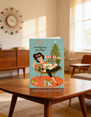 A holiday greeting card featuring a woman sitting in a chair surrounded by wrapped gifts, with a Christmas tree in the background.