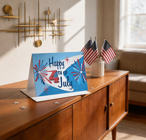 A greeting card with the text "Happy 4th of July" sits on a wooden table, surrounded by American flags and a decorative gold wall hanging.