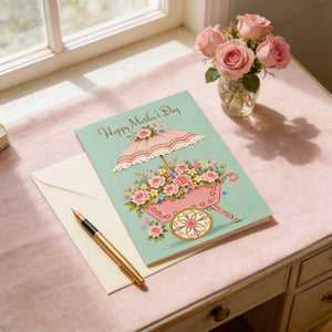 A Mother's Day card with a pink umbrella and flowers sits on a pink table next to a vase of pink roses, with a gold pen and a window in the background.