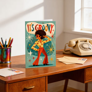 A colorful greeting card with a dancing man and the text "It's groovy" is displayed on a wooden desk, accompanied by a telephone, a pen holder, and some envelopes.