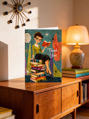 A greeting card with an illustration of a woman sitting on a stack of books, reading a book, and surrounded by books, with a lamp and a wall decoration in the background.