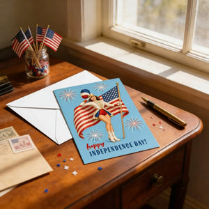 A desk with a card featuring a woman holding an American flag, surrounded by fireworks, and a pen, along with a jar of American flags and a window.