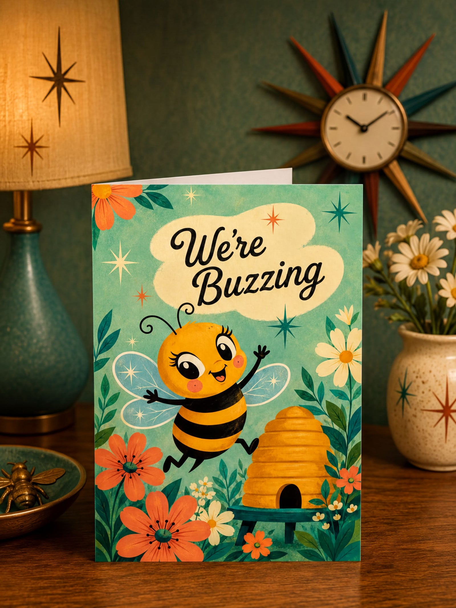 A folded greeting card shown front-facing on a white background. The card features a cute cartoon bee with the text 'We're Buzzing' above it, surrounded by flowers, leaves, stars, and a beehive.