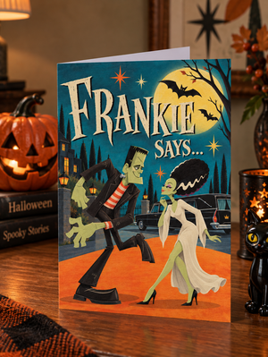 A Halloween greeting card displayed on a wooden table, featuring an illustration of Frankenstein's monster and Dracula under a full moon with the text 'FRANKIE SAYS.