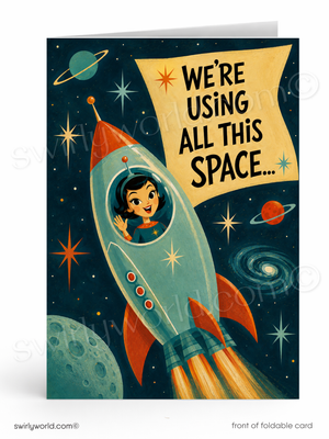 A foldable greeting card shown upright on a white background. The card front features a retro space illustration with a teal rocket carrying a child astronaut, planets, stars, and a yellow banner that reads, 'WE'RE USING ALL THIS SPACE.