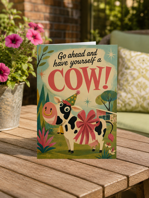 A square greeting card featuring a whimsical illustration of a cow wearing a party hat and a large pink bow, with the text 'Go ahead and have yourself a COW!' on the front. The card is displayed standing upright on a wooden table in an outdoor garden setting with flowers and a sofa in the background.