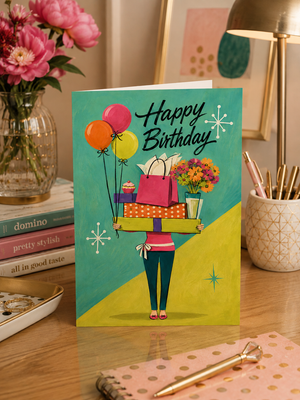 A colorful 'Happy Birthday' greeting card featuring an illustration of a person carrying gift bags, balloons, and flowers, displayed on a wooden desk surrounded by stationery and flowers.