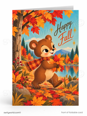 A foldable greeting card shown standing upright, featuring a whimsical autumn illustration of a brown bear holding orange leaves near a tree with colorful fall foliage, a lake, mountains, and the words “Happy Fall.”.