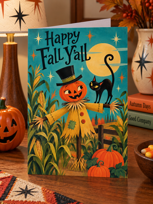 A Halloween greeting card standing on a wooden table, featuring a smiling pumpkin scarecrow, a black cat, corn, pumpkins, and the text 'Happy Fall Y'all' against a teal sky with a yellow moon. The card is styled in a cozy autumn setting with a small jack-o'-lantern, books, and decorative vases.