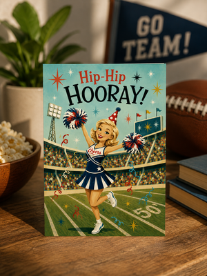 A rectangular greeting card featuring a retro-style illustration of a cheerleader on a football field with the text 'Hip-Hip HOORAY!' stands on a wooden table. The card is styled in a cozy indoor setting with a plant, popcorn bowl, football, and books in the background.