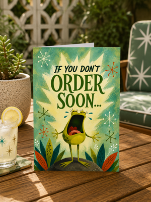 A greeting card standing on a wooden table outdoors, featuring a colorful illustration of a crying avocado with the text 'IF YOU DON'T ORDER SOON.