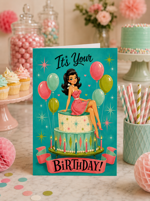 A retro-style birthday card featuring a pin-up illustration of a woman sitting on a tiered cake with balloons and the text 'It's Your Birthday!' The card is displayed on a marble surface in a decorated party setting with cupcakes, candy jars, and pastel props.