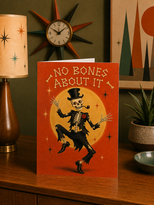 A greeting card featuring a skeleton dancing in front of a yellow moon with the text 'NO BONES ABOUT IT' on an orange-red background, styled on a wooden table with a lamp, plant, and wall decor in the background.
