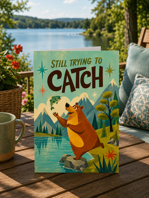 A greeting card featuring a cartoon brown bear trying to catch a fish over a lake, styled on a wooden deck with a mug and outdoor scenery in the background.