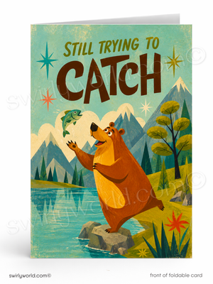 A folded greeting card shown standing upright, featuring a retro-style illustration of a brown bear trying to catch a fish over a lake with mountains and trees in the background. The card text reads 'STILL TRYING TO CATCH'.