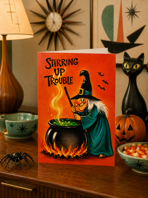 A Halloween greeting card standing on a wooden table, featuring an illustration of a witch stirring a cauldron with the text 'Stirring Up Trouble'. The card is styled in a cozy, decorated room with a lamp, a black cat figurine, a pumpkin, and a bowl of candy.