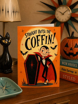 A Halloween-themed greeting card featuring a cartoon vampire standing next to a coffin with the text 'STRAIGHT OUTTA THE COFFIN!' on the front. The card is displayed on a wooden table in a styled room setting with a black cat figurine, a starburst clock, a jack-o'-lantern, and books in the background.