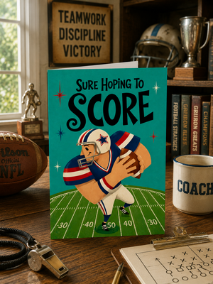 A football-themed greeting card featuring an illustration of a quarterback on a teal field with the text 'SURE HOPING TO SCORE', displayed on a wooden desk surrounded by sports memorabilia.