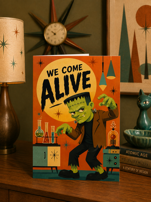 A retro-style Frankenstein greeting card standing on a wooden table, surrounded by vintage decor including a lamp, star clock, framed art, and a small cat figurine. The card features an illustrated green monster with the text 'WE COME ALIVE'.